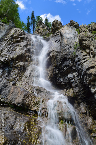 Shirlak waterfall in rocks on blue sky with white clouds background. Tektu river, Altai Mountains, Altay Republic, Siberia, Russia. Vertical orientation.