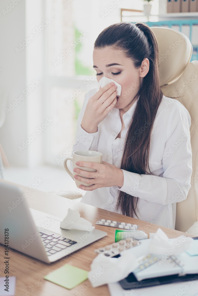 Young ill business woman economist is sneezing at workplace in modern office, with cup of coffee, a lot of pills on desktop Young ill business woman economist is sneezing at workplace in modern office, with cup of coffee, a lot of pills on desktop