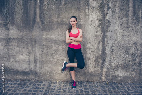 Young serious slim trainer is stretching her leg by doing exercise. She is training outdoors on a summer day, wearing fashionable sport wear, sneakers, with pony tail