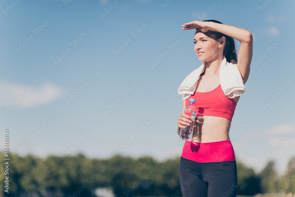 Young fit sportwoman finished her work out and now drinking water and smiling. She is outside on a summer stadium, with a towel on her shoulders