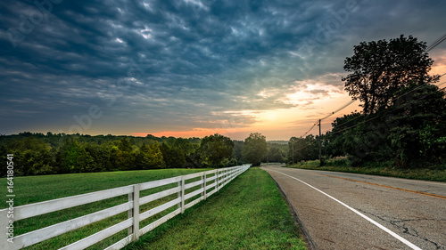 A white wooden fence between a country meadow and blacktop road at sunrise