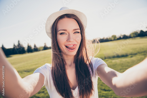 Playful funky selfie mood! Happy young girl on vacation in an open air spring park is making selfie, wearing casual outfit, hat, posing with tongue out © deagreez