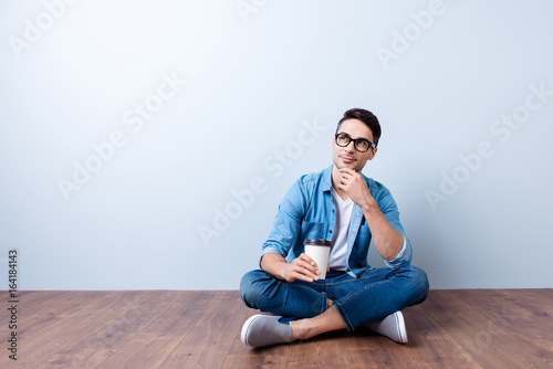 Happy student is sitting on the floor at home with crossed legs, relaxing with cup of tea. He is in a casual outfit, glasses, dreamy and pensive © deagreez