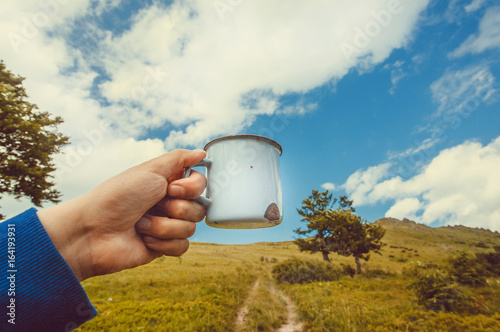 Mug of water or hot tea in hand of a lonely tourist. Hike concept in green grass mountains with dirt road.
