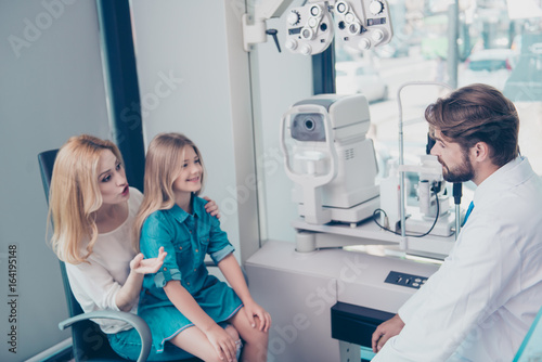 Optometrist consultation. Blond mom with cheerful girl are talking to brunet bearded male doctor optician in a hospital