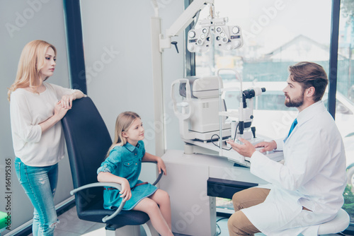Optometrist consultation. Blond mom with smiley girl are talking to brunet bearded male doctor optician in his office about the diagnosis of the small patient