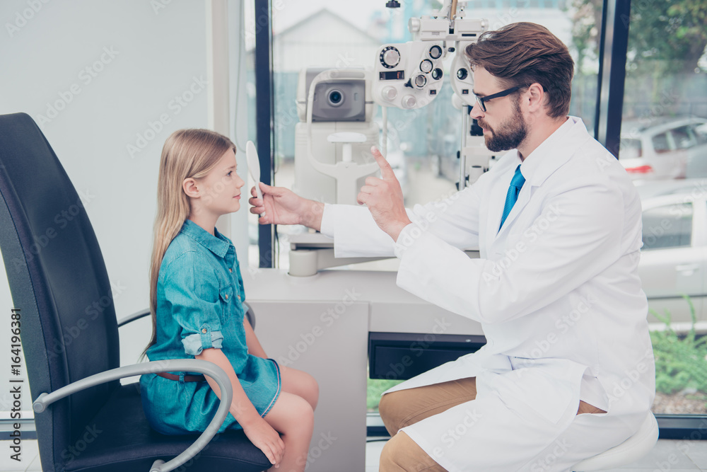 Fototapeta premium Health care, medicine, eye sight and technology concept. Side profile of brunet bearded optometrist is checking small blond`s girl patient eye sight at his office