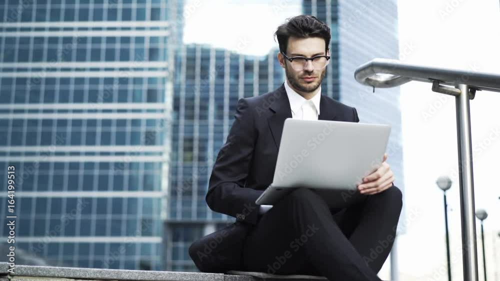 Front view of a handsome young businessman wearing glasses and a suit sitting on the stairs and working with his laptop. Handheld real time medium shot Front view of a handsome young businessman wearing glasses and a suit sitting on the stairs and working with his laptop. Handheld real time medium shot