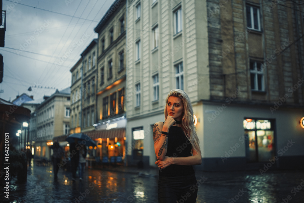 Young woman with tattoo on shoulder stands on city street in evening in rain.