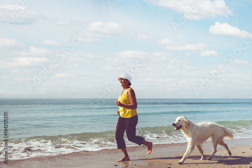 Happy elderly woman running along a beach with her golden retriever at the morning