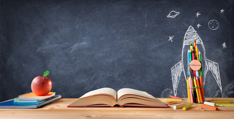 Start School Concept - Supplies On Desk And Rocket Drawn On Blackboard
