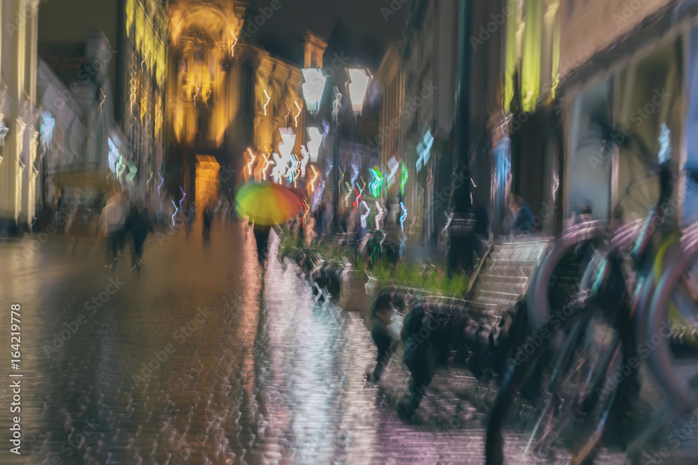 Abstract blurred background of girl under colorful vivid umbrella, city street in rain. Light illumination from lanterns, shop windows. Impressionism style, blur. Modern city, lifestyle, leisure.