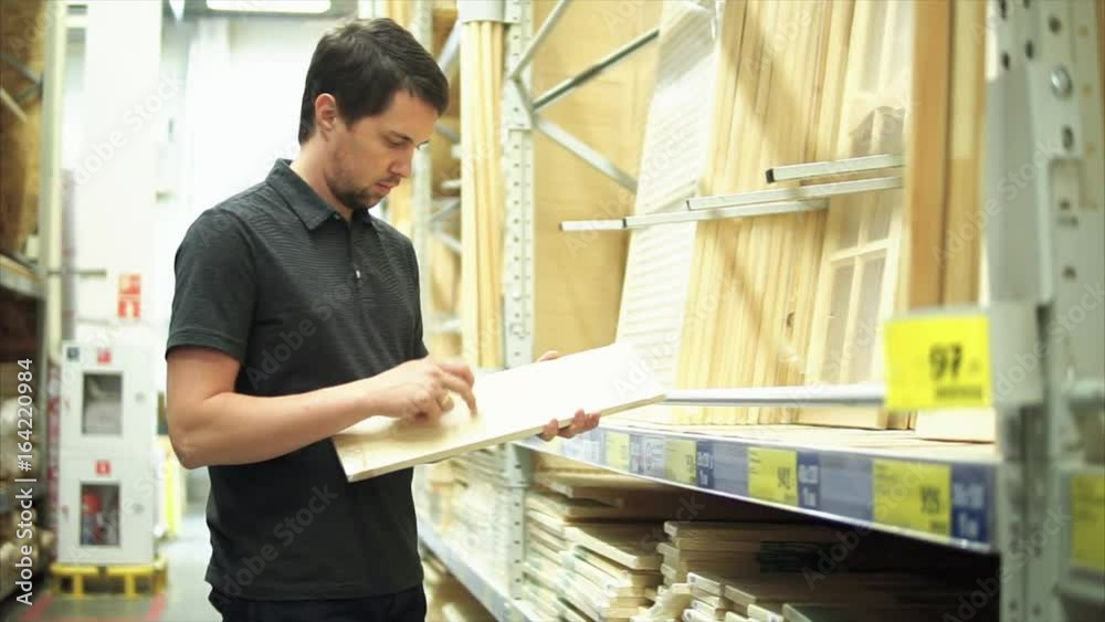 Male customer in the hardware store standing near shelf of wooden crafts. He taking the wooden plank and knocking on it. Male customer in the hardware store standing near shelf of wooden crafts. He taking the wooden plank and knocking on it.