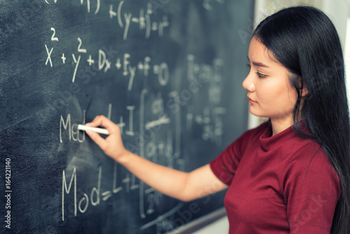 Beautiful Asian student writing on blackboard with chalk in classroom. photo