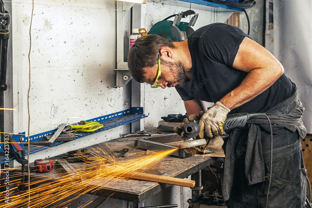 A young man welder in a black T-shirt, glasses and construction gloves grinder metal an angle grinder in the garage, in the background a lot of tools, sparks fly to the side A young man welder in a black T-shirt, glasses and construction gloves grinder metal an angle grinder in the garage, in the background a lot of tools, sparks fly to the side