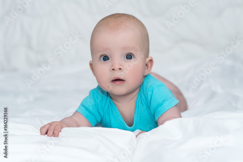 Adorable baby girl or boy in blue shirt with big blue eyes lying on her stomach on soft blanket and looks with surprised, indoors