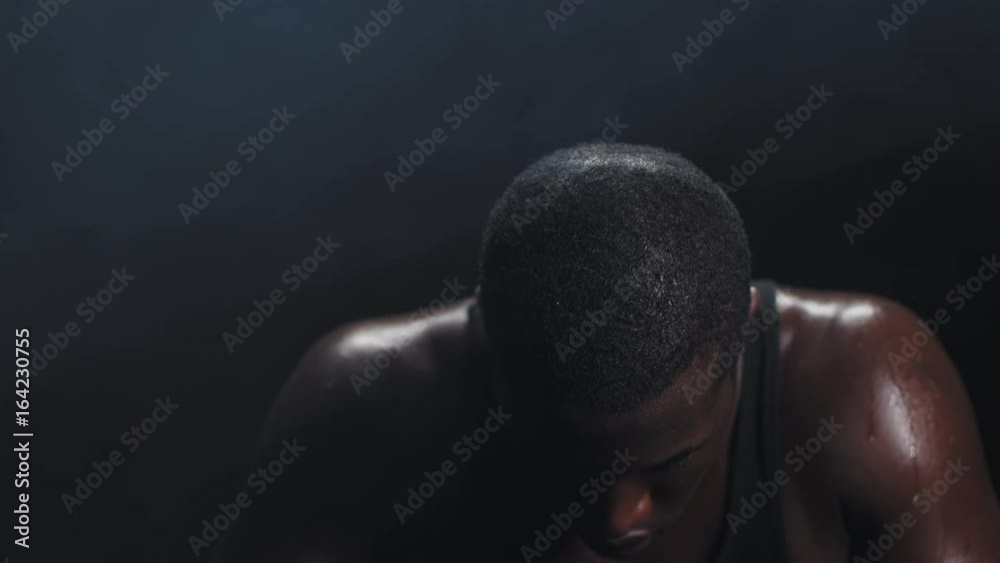 Zooming shot of a muscular black man wiping the sweat off his forehead on a foggy dark background