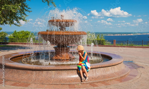 little girl is sitting near fountain on waterfront on summer day photo