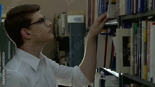 A young man with glasses, dressed in a white shirt, searches for a book in the library. photo