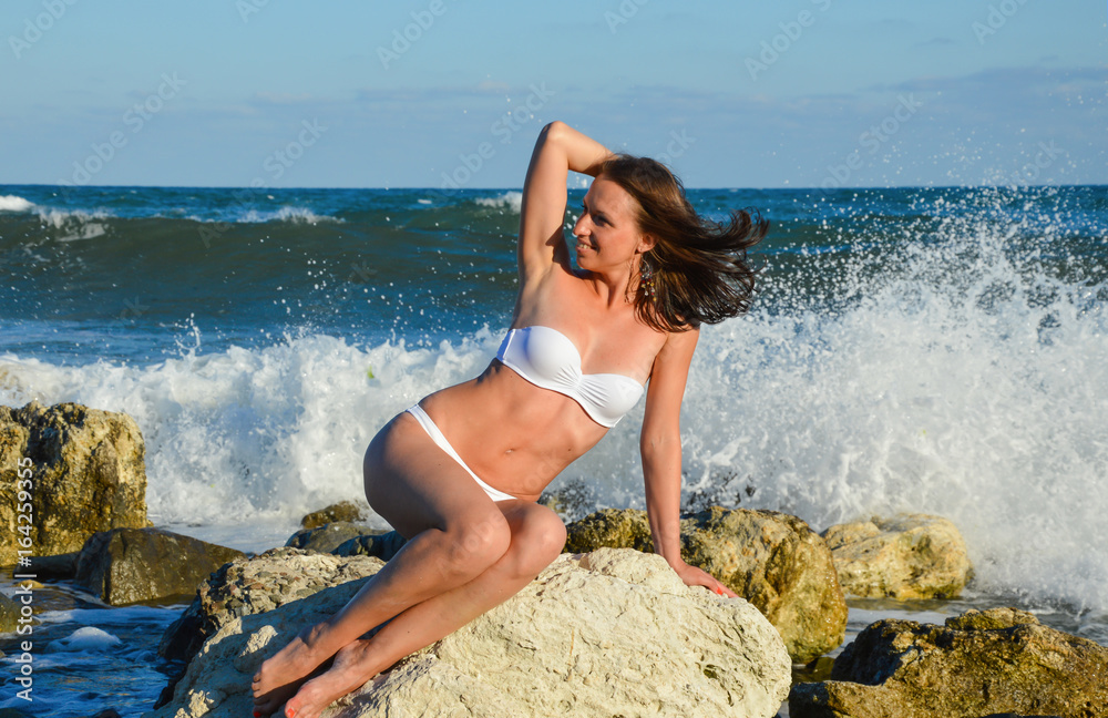 Sexy young brunette beauty wearing white swimsuit sitting on stone at the beach over beautiful sea