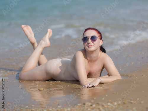 Naked red-haired girl in sunglasses tans on wet sand