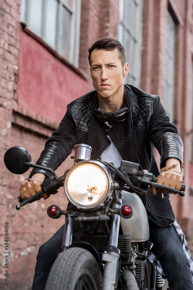 Handsome rider biker man in black leather jacket, jeans, boots and gloves  sit on classic style cafe racer motorcycle. Bike custom made in vintage  garage. Brutal fun urban lifestyle. Outdoor portrait. Stock