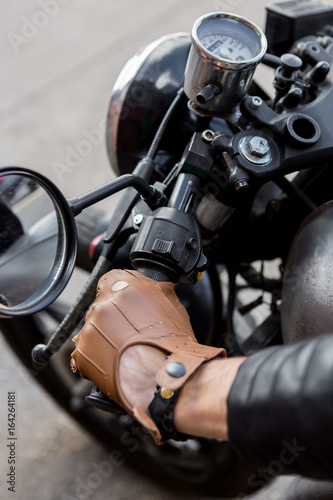 Close up of a hipster biker guy hand in leather glove hold throttle control of classic style cafe racer motorcycle. Bike custom made in vintage garage. Brutal fun urban lifestyle. Outdoor portrait. © Mikalai Bachkou