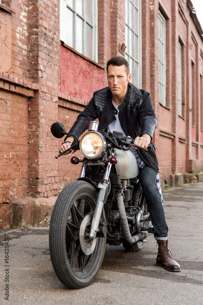 Handsome rider biker guy in leather jacket sit on classic style cafe racer motorcycle and ready for long ride. Bike custom made in vintage garage. Brutal fun urban lifestyle. Outdoor portrait.