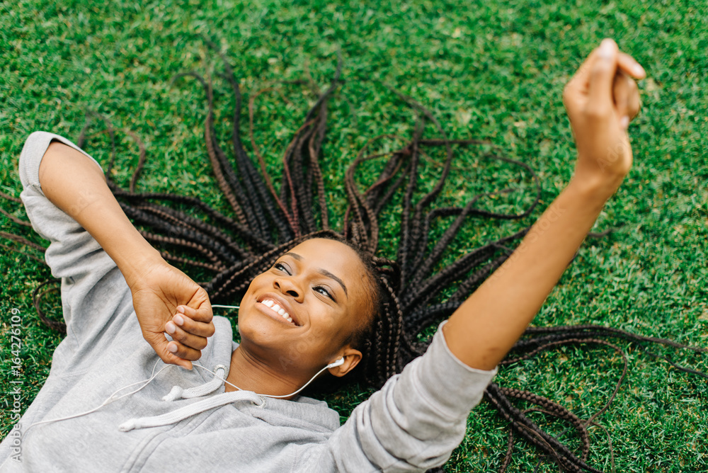 Fototapeta premium Above portrait of the joyful afro-american teenager laying on the grass while listening to music and moving hands.