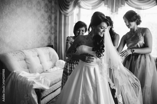 Stylish beautiful bridesmaids helping gorgeous brunette bride in white dress get ready for wedding, morning preparations, woman putting on dress