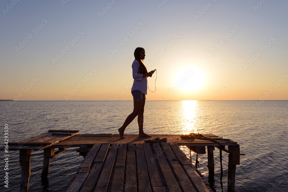 Happy cheerful young girl in a white shirt listening to music on headphones and dancing on the dock by the sea at sunset