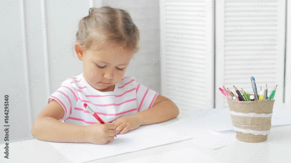 Cute preschool girl sitting by the white table focused on drawing something. Cute preschool girl sitting by the white table focused on drawing something.