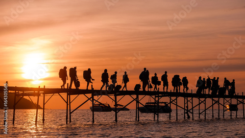 Silhouettes of the backpackers walking along the wooden pier to the ferry on beautiful sunset time. Tourists with bags walk along the Bang Rack pier to the ferry to Phangan island photo
