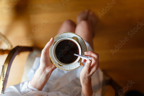 Morning coffee mug in the girl's hands in my nightie top view. Happy young woman in a white Bathrobe drinking a Cup while enjoying the morning sitting in a wooden chair.
