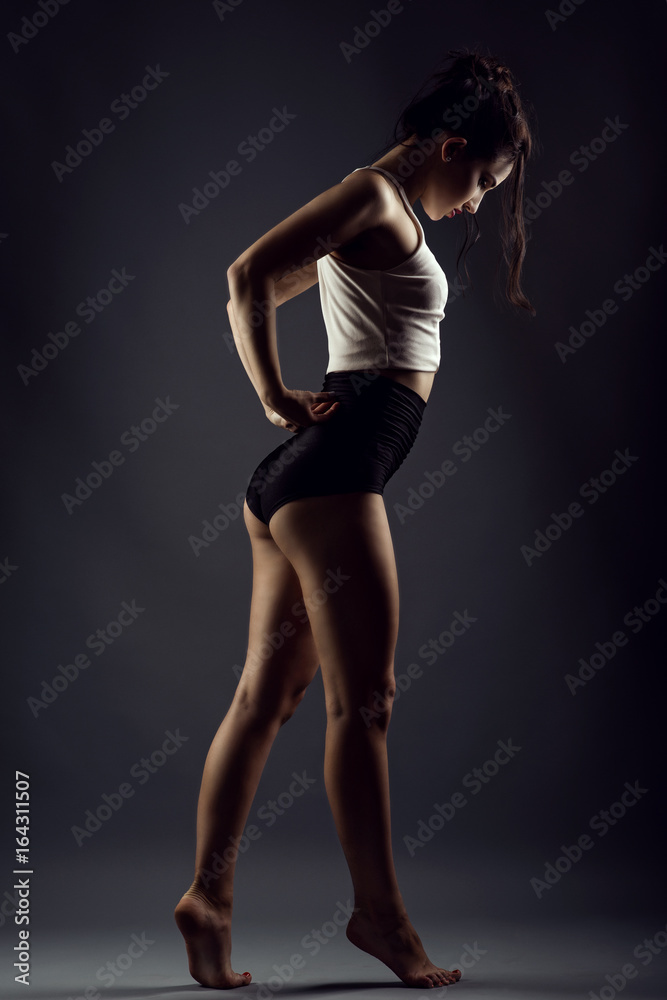 Portrait of a beautiful slender girl athlete with updo hair dancing barefoot under studio searchlights wearing black high waist panties and white top. Isolated on dark background