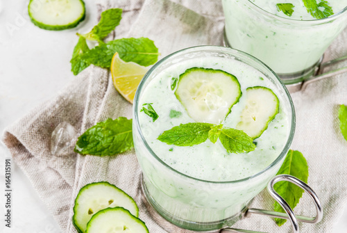 Summer food. Refreshing dishes. Cold soup of cucumber, avocado, with herbs and mint. With serving glasses, with slices of cucumber. On a white concrete table, with ingredients. Copy space photo