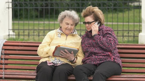 Two women sit and discuss about home problems outdoors. One woman aged 80s dressed in yellow jacket holds computer tablet another woman talking with friend using a smart phone photo