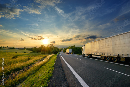 Obraz na płótnie Trucks driving towards the setting sun on a tarmac road in rural countryside w salonie
