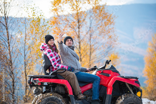 Close-up of guy with girlfriend in winter clothing on red quad bike makes selfie on the phone against blurred background of landscape mountains and trees with yellow leaves