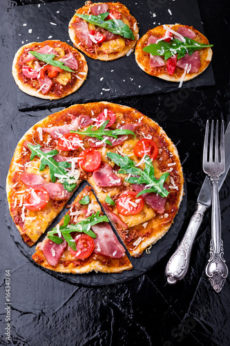 Homemade pizza with prosciutto, tomato, arugula on black slate board. Top view.