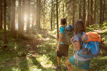 Man and woman with backpack walking on hiking trail path in forest woods during sunny day.Group of friends people summer adventure journey in mountain nature outdoors.Travel exploring Alps,Dolomites
