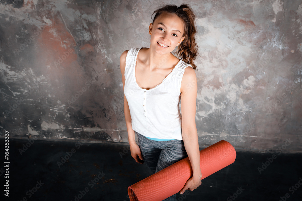 Sim fitness young beautiful woman standing, top view, on gray background, holding pink yoga mat, indoor, on gray background, upper half, loft, urban style, looking camera Sim fitness young beautiful woman standing, top view, on gray background, holding pink yoga mat, indoor, on gray background, upper half, loft, urban style, looking camera