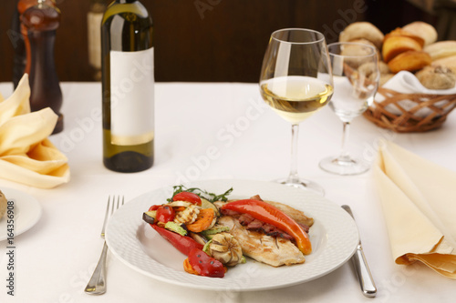 Chicken with bacon and grilled vegetables arranged on a plate  Wine bottle and wineglass in background  Traditional dish in elegant setting  Selective focus with soft light