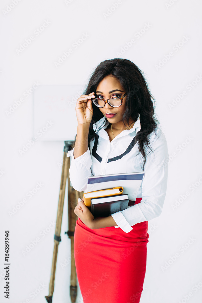 Beautiful young african smiling woman teacher holding books and documents near the bord