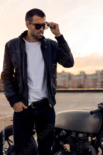 Close up of a handsome rider guy with beard and mustache in black biker jacket and sunglasses smoking cigaret near classic style cafe racer motorbike on rooftop at sunset. Brutal fun urban lifestyle.