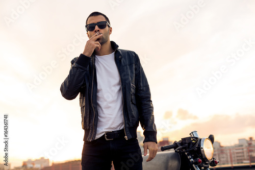 Beautiful happy rider guy with beard and mustache in black biker jacket, jeans and fashion sunglasses smoking cigaret near classic style cafe racer motorbike at sunset. Brutal fun urban lifestyle.