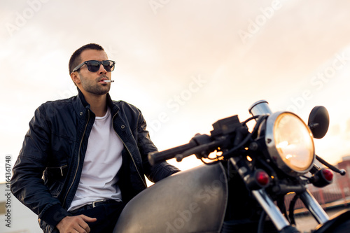 Beautiful happy rider man with beard and mustache in black biker jacket, denim and fashion sunglasses smoking cigaret sit on classic style cafe racer motorbike at sunset. Brutal fun urban lifestyle.