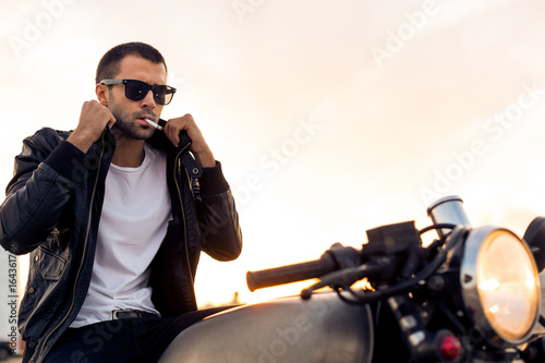 Beautiful happy rider man with beard and mustache in black fashion sunglasses smoking cigaret and correct biker jacket sit on classic style cafe racer motorbike at sunset. Brutal fun urban lifestyle.