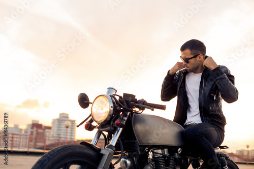 Handsome rider man with beard and mustache in black fashion sunglasses smoking cigaret and correct biker jacket sit on classic style cafe racer motorbike at sunset. Brutal fun urban lifestyle.