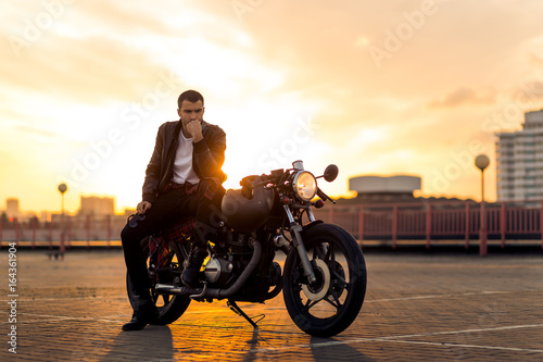 Handsome fit rider guy with beard and mustache in black leather biker jacket sit on classic style cafe racer motorcycle at sunset time. Bike custom made in vintage garage. Brutal fun urban lifestyle.